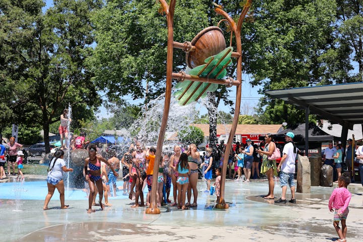 A group of people, mostly children, enjoying a splash pad on a sunny day, surrounded by trees and onlookers.