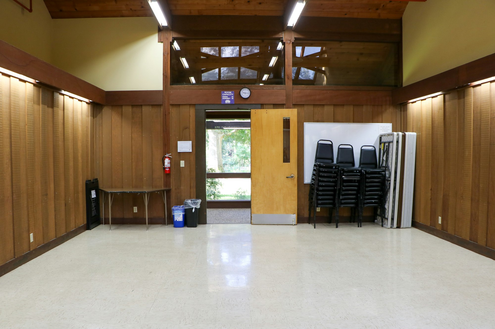An empty room with wood paneling, chairs stacked, a door, clock, recycle bin, and a fire extinguisher.