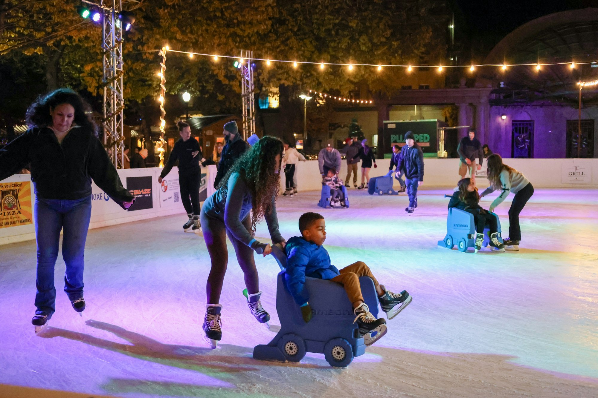People ice skating at an outdoor rink, some using aids, under string lights at night.