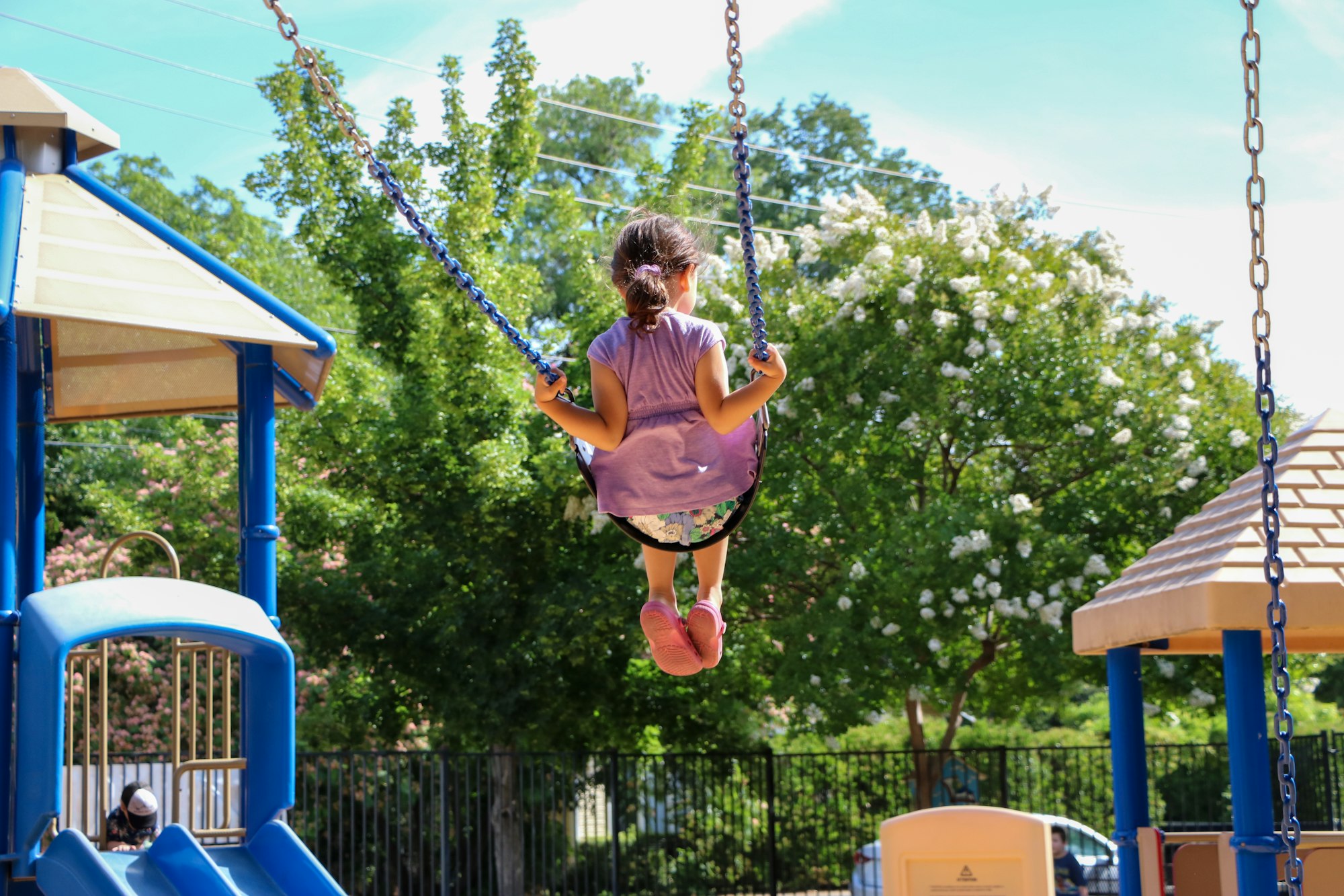 A child swinging at a playground surrounded by trees and play structures under a sunny sky.