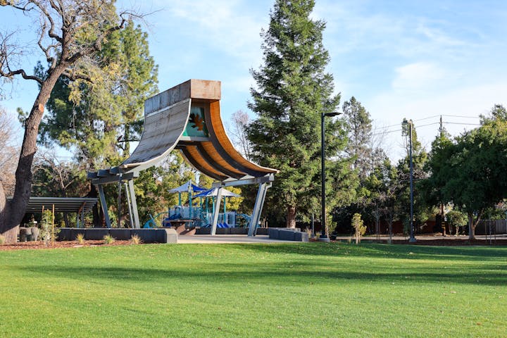 A playground with blue equipment and a unique canopy set in a park with green grass and trees.