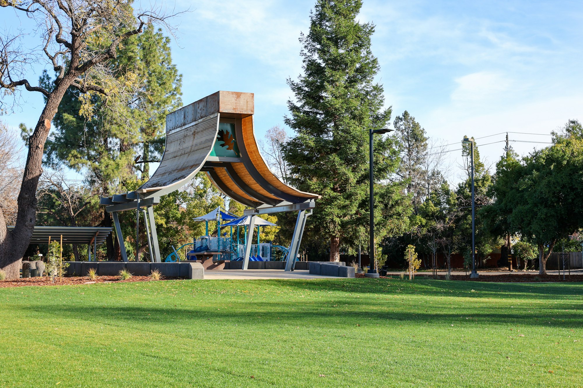 A playground with blue equipment and a unique canopy set in a park with green grass and trees.