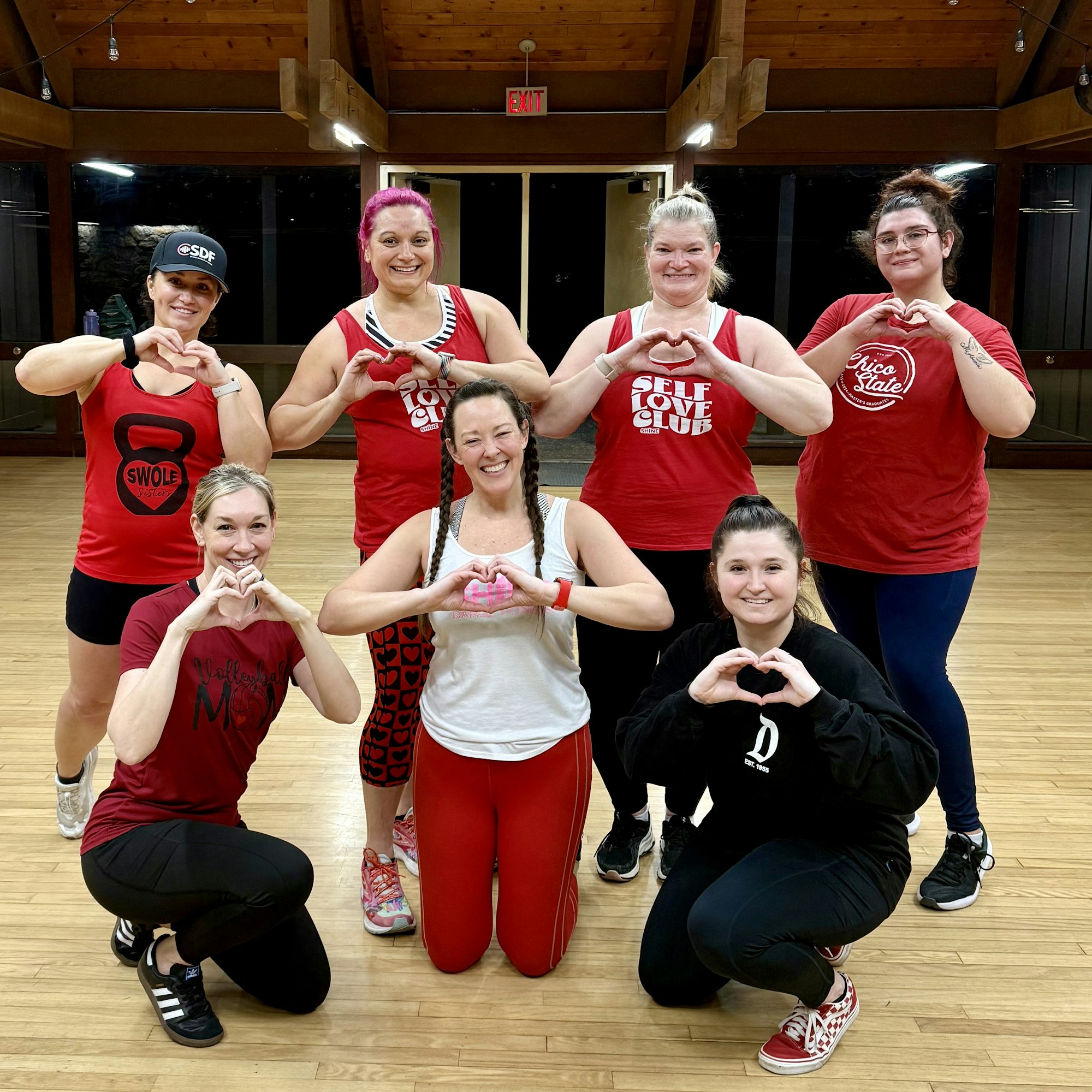 Eight people in gym attire posing indoors, forming heart shapes with their hands.