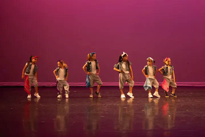 A group of young girls performing a dance on stage, dressed in casual outfits with bandanas against a purple backdrop.
