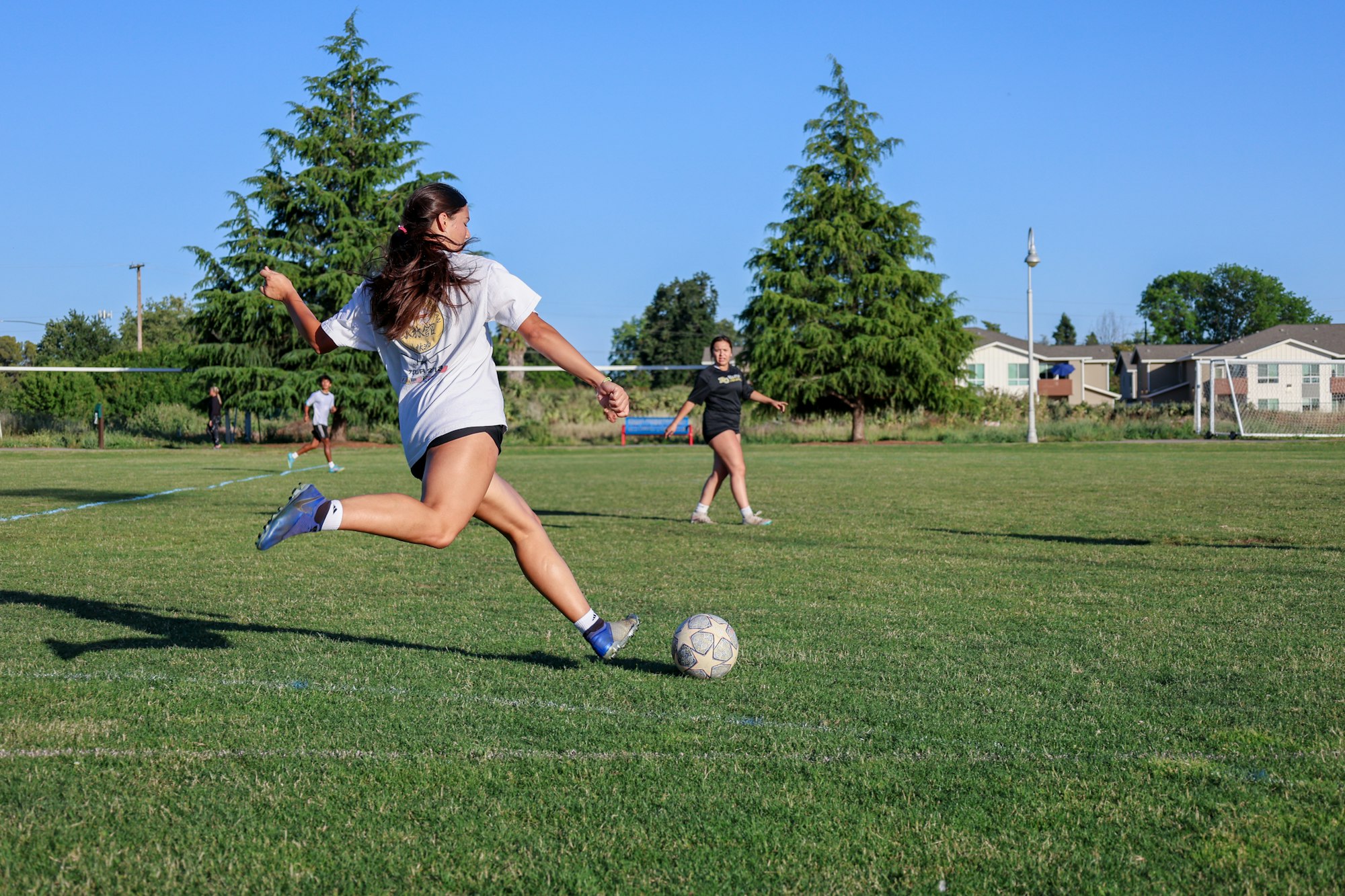 A person kicking a soccer ball on a sunny field with others in the background.