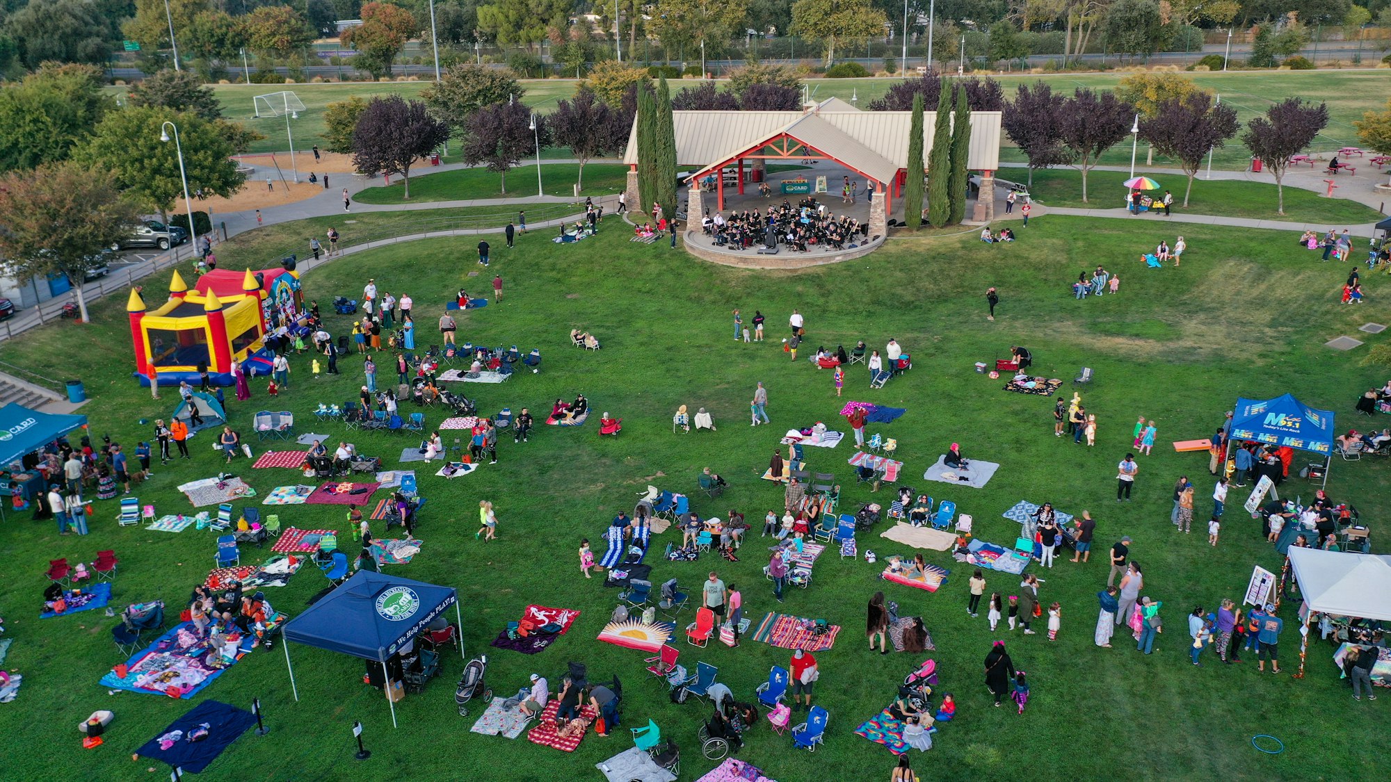A community event in a park with people on blankets, a bounce house, and a band playing under a pavilion.