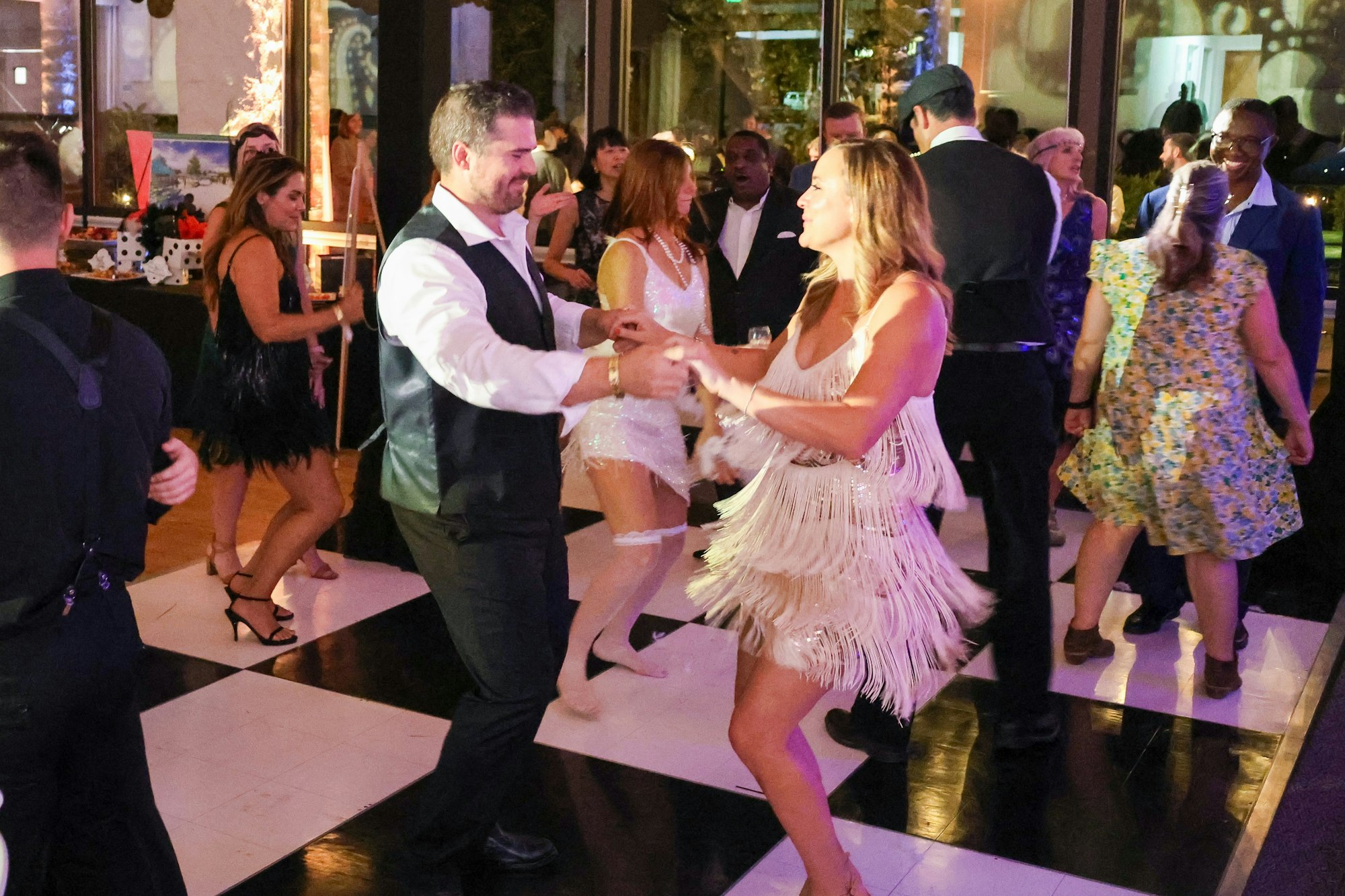 People dancing at a party on a checkered floor, wearing festive attire.
