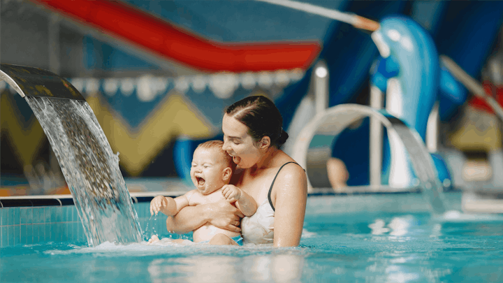 Happy mother holding a smiling baby in a pool at an aquatics center.