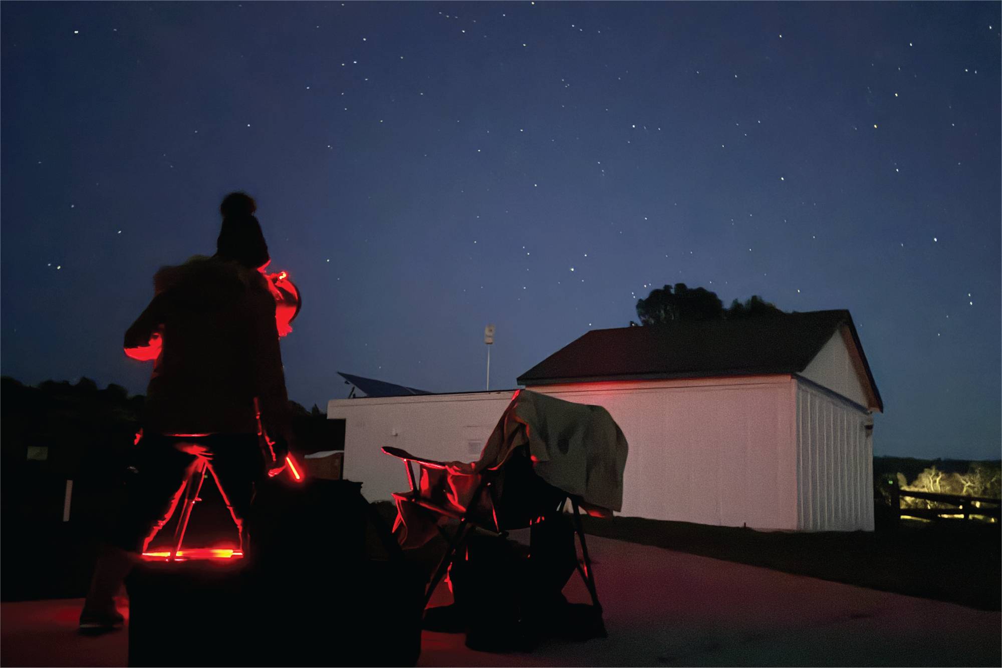 Silhouetted person stargazing with telescopes at night under a starry sky, next to a building.