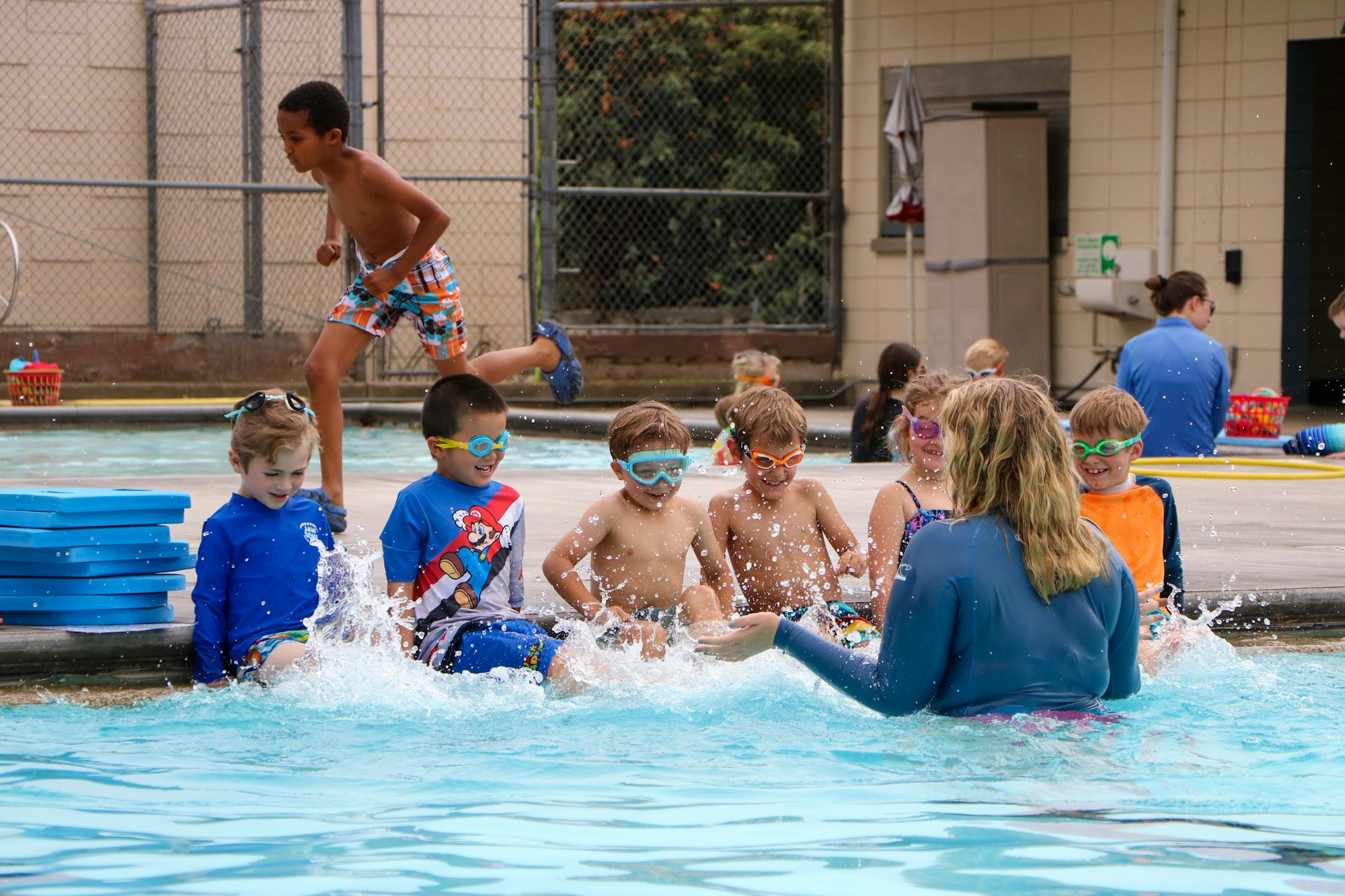 Children with swimming gear splash in a pool under adult supervision, while one boy leaps into the water.