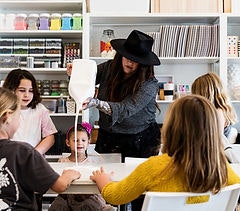 A woman in a hat is pouring liquid into a container while several kids watch and engage in a creative activity.
