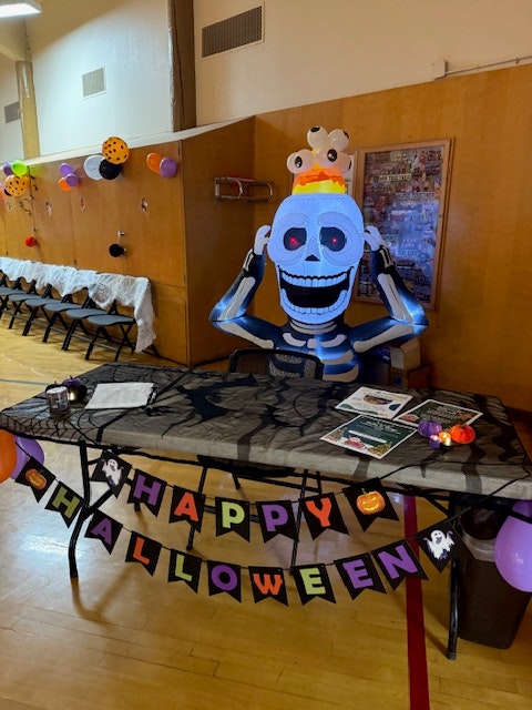 A decorated table for Halloween featuring a large skeleton figure, festive banners, and themed decorations like balloons and spider webs.