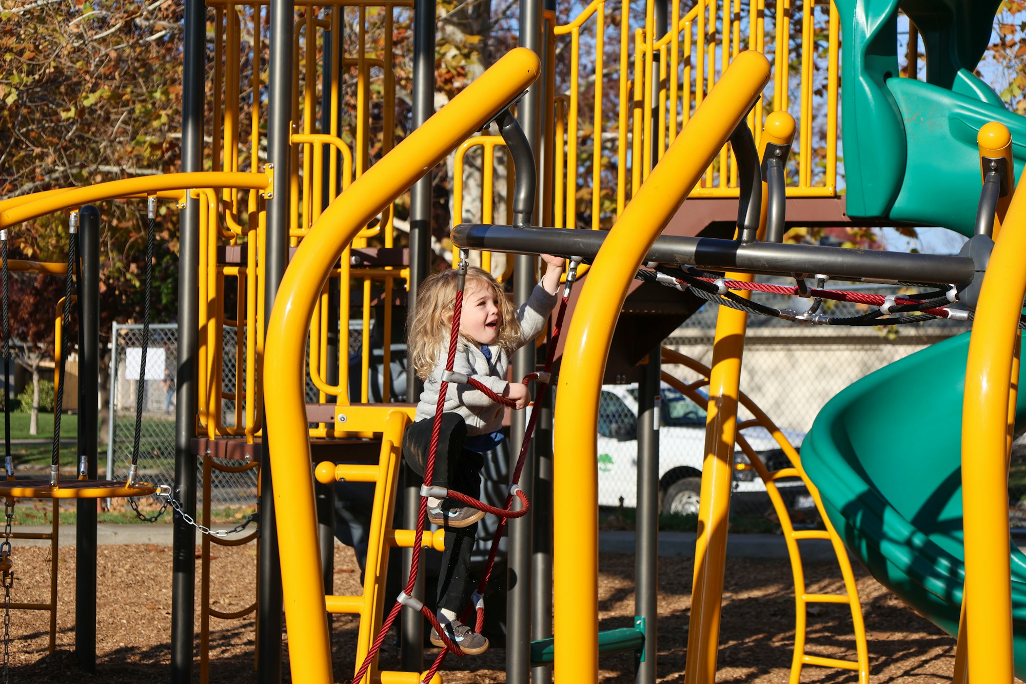 A child with curly hair is playing on a yellow and green playground structure, smiling while climbing.