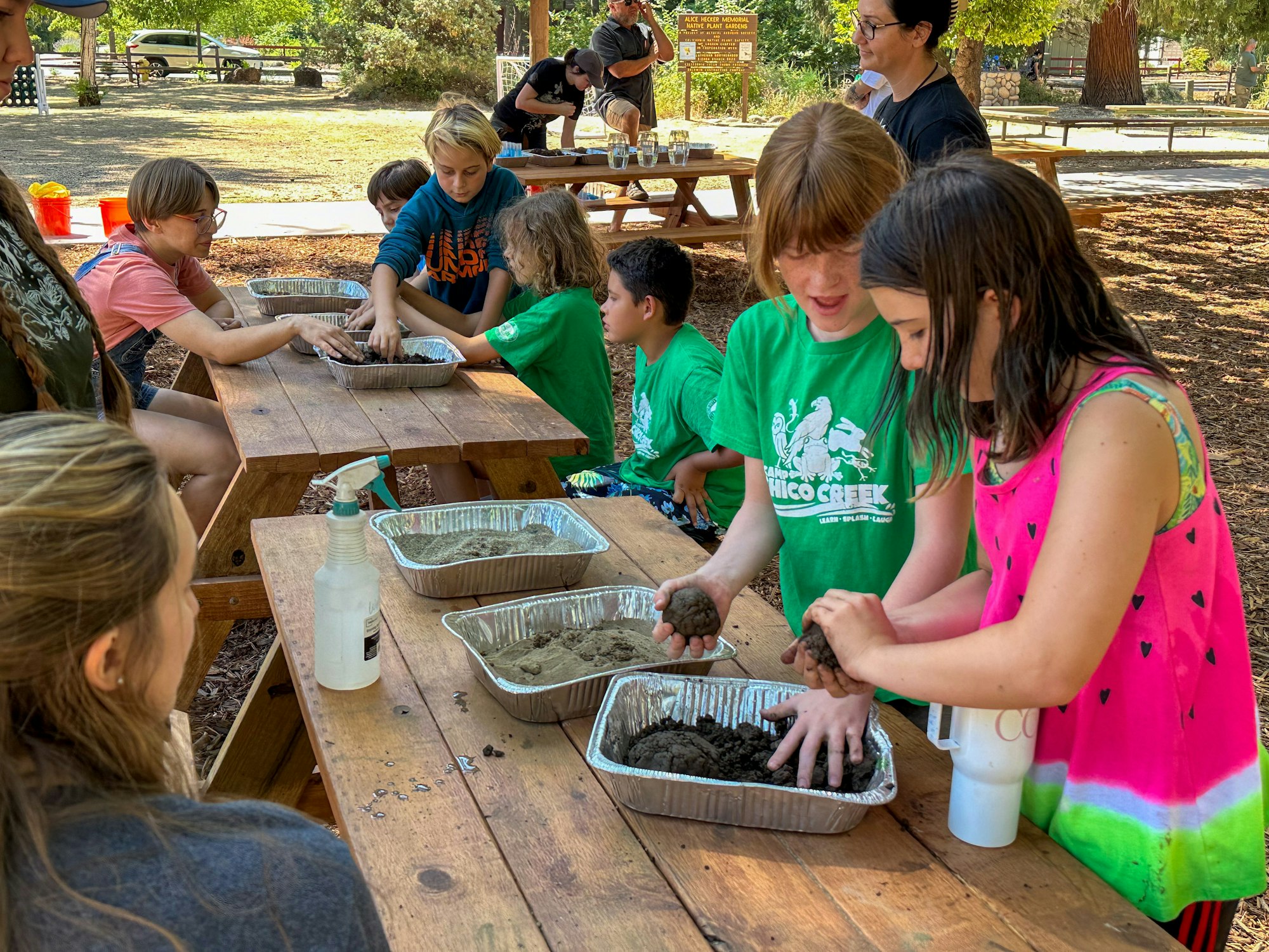 Children outdoors making mud projects at picnic tables, supervised by adults.