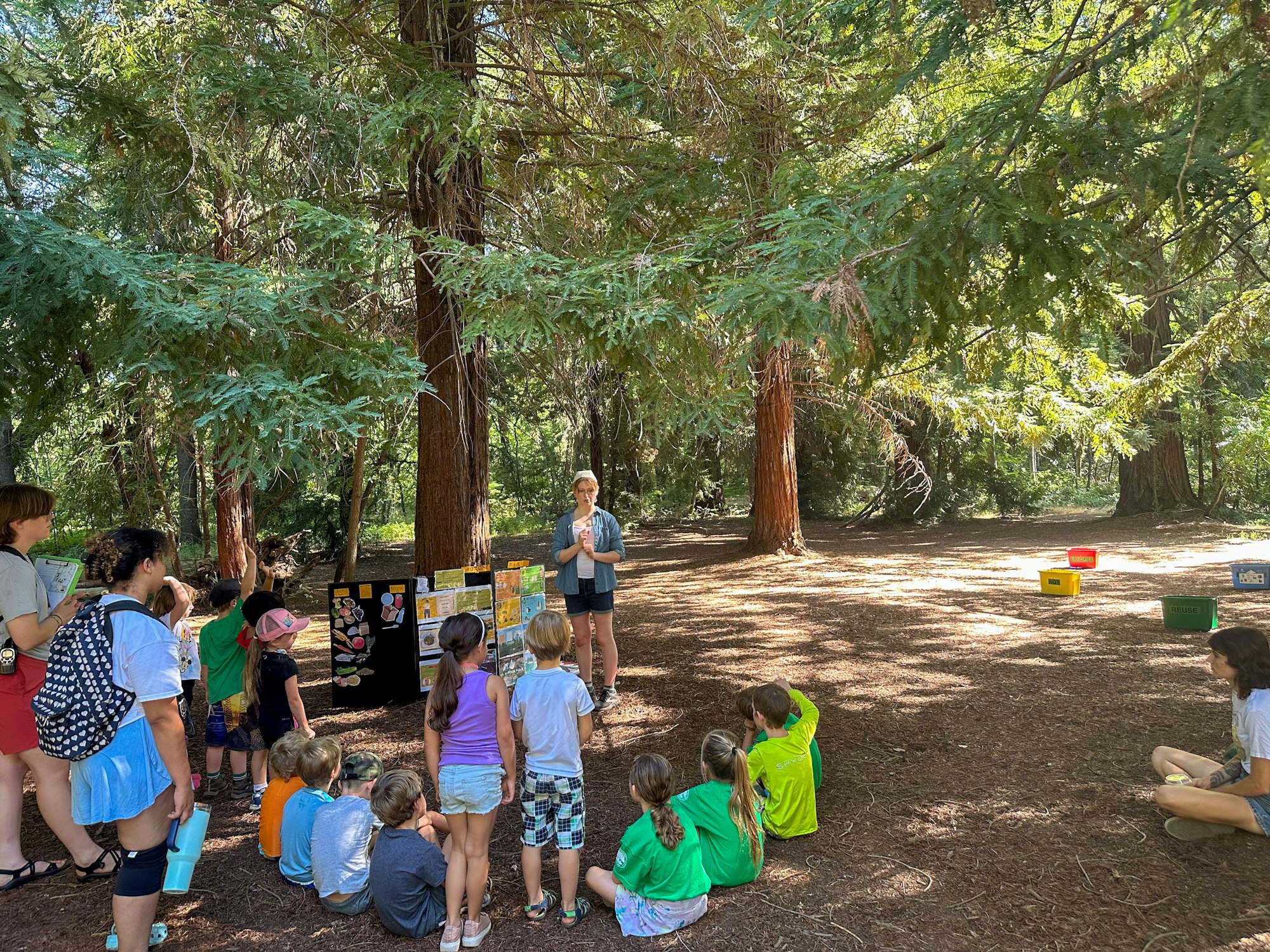 A group of kids and adults are outdoors, sitting and standing, engaged in an educational session with posters about nature.