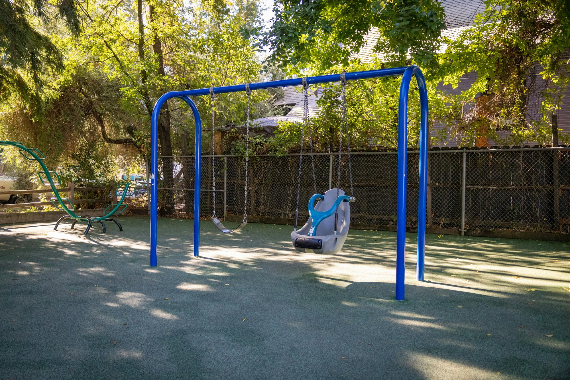 A playground swing set with a blue frame and a child-safe swing, surrounded by trees and a soft rubber surface.