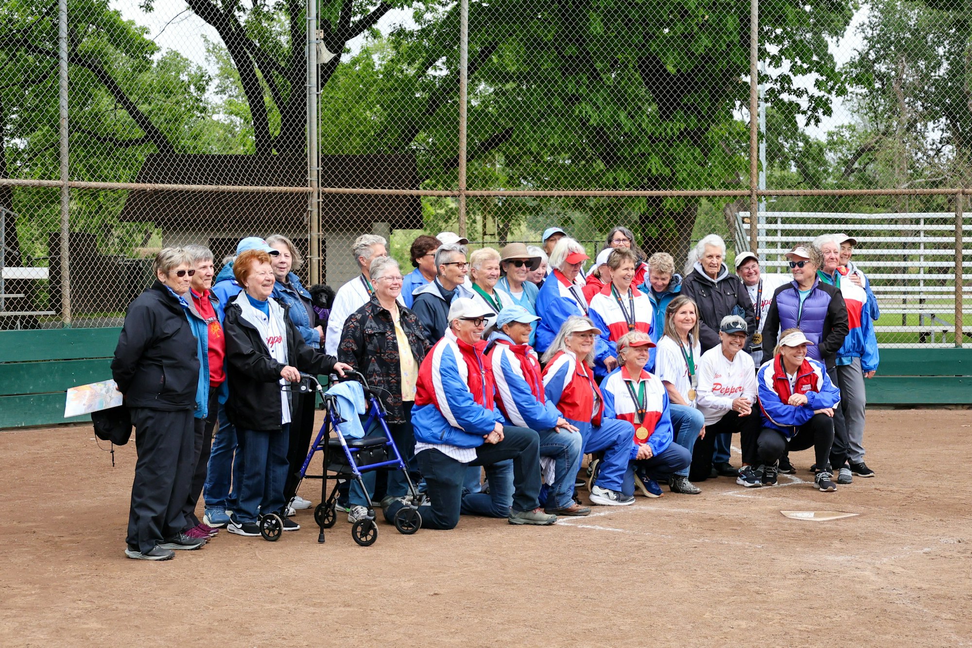 Group of people posing on a baseball field, some kneeling, wearing jackets and medals, with trees and a fence in the background.