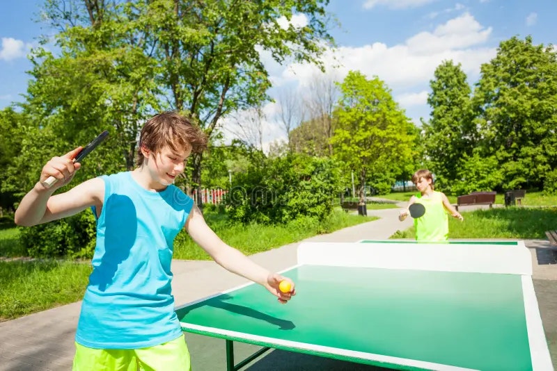 Two boys are playing table tennis outdoors, enjoying a sunny day in a park. One is serving while the other is ready to return.