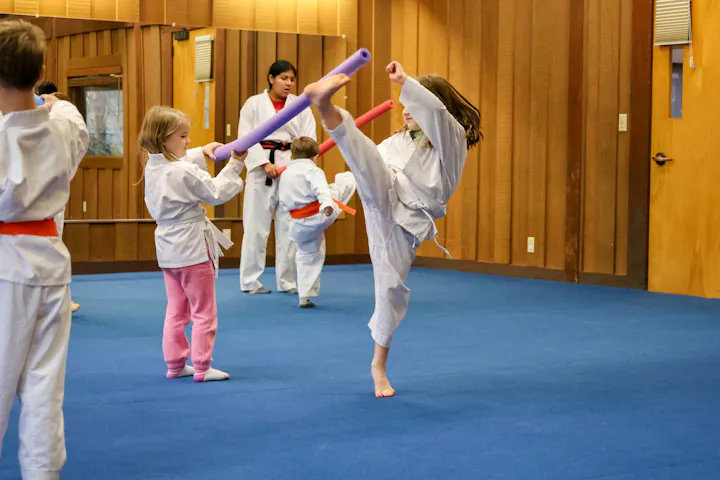 Children in karate uniforms practice martial arts with colorful foam noodles in a dojo setting.