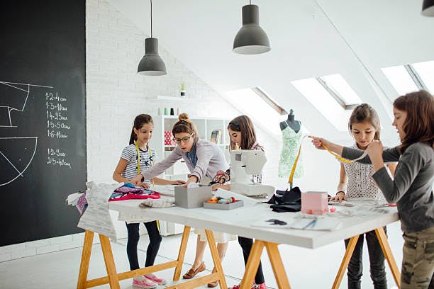 A sewing workshop with one adult and four kids working on fabrics with a sewing machine and materials on the table.