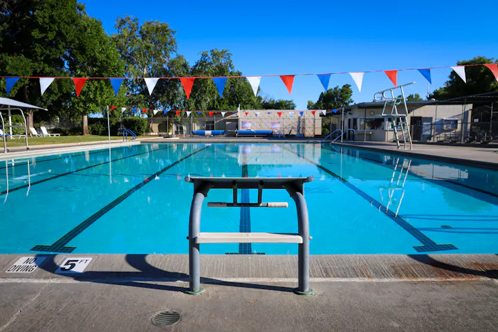 Outdoor swimming pool with flags and starting block, surrounded by trees and buildings, under a clear blue sky.