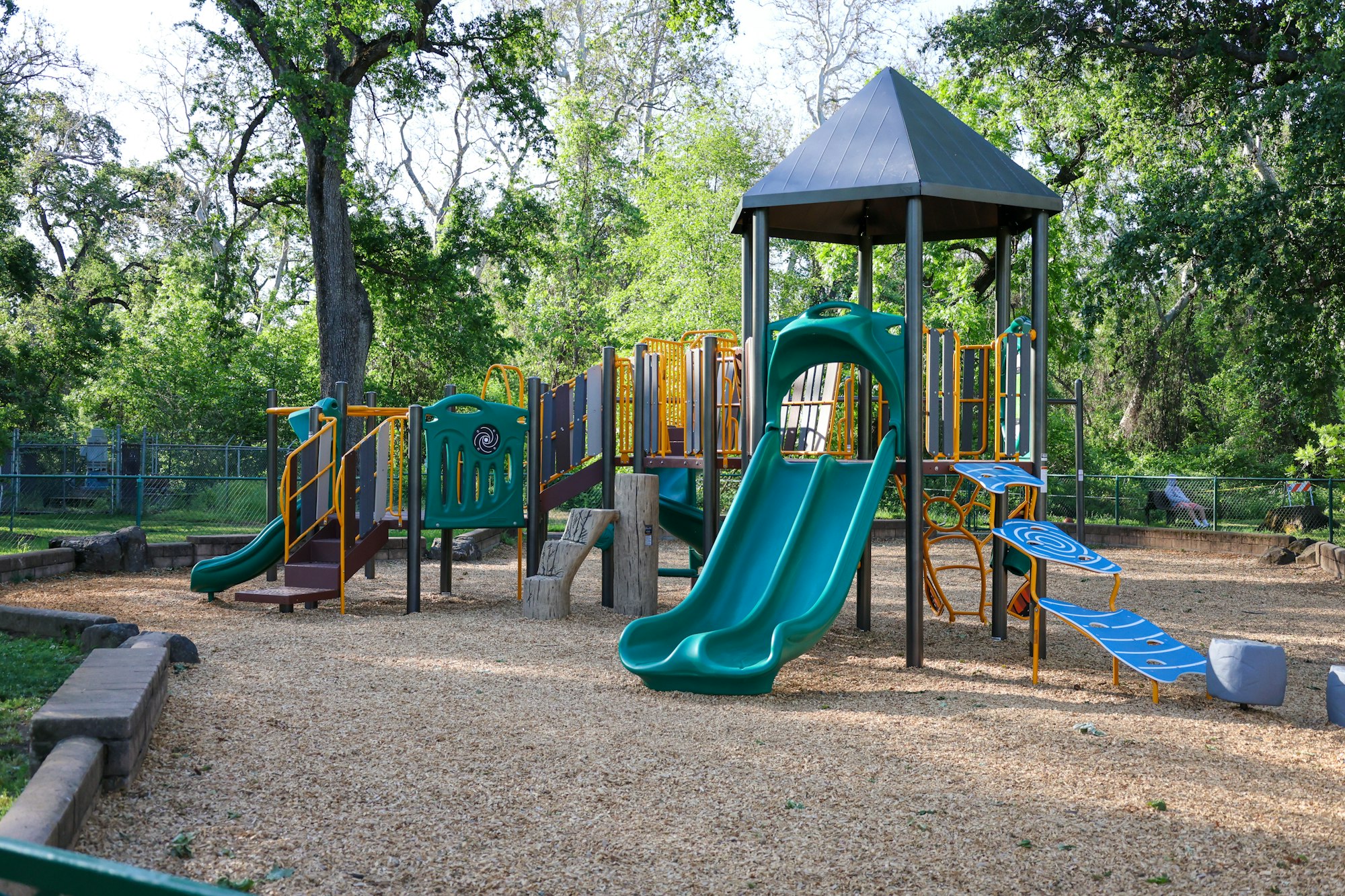 A colorful playground with slides and climbing structures, surrounded by trees and wood chips on the ground.