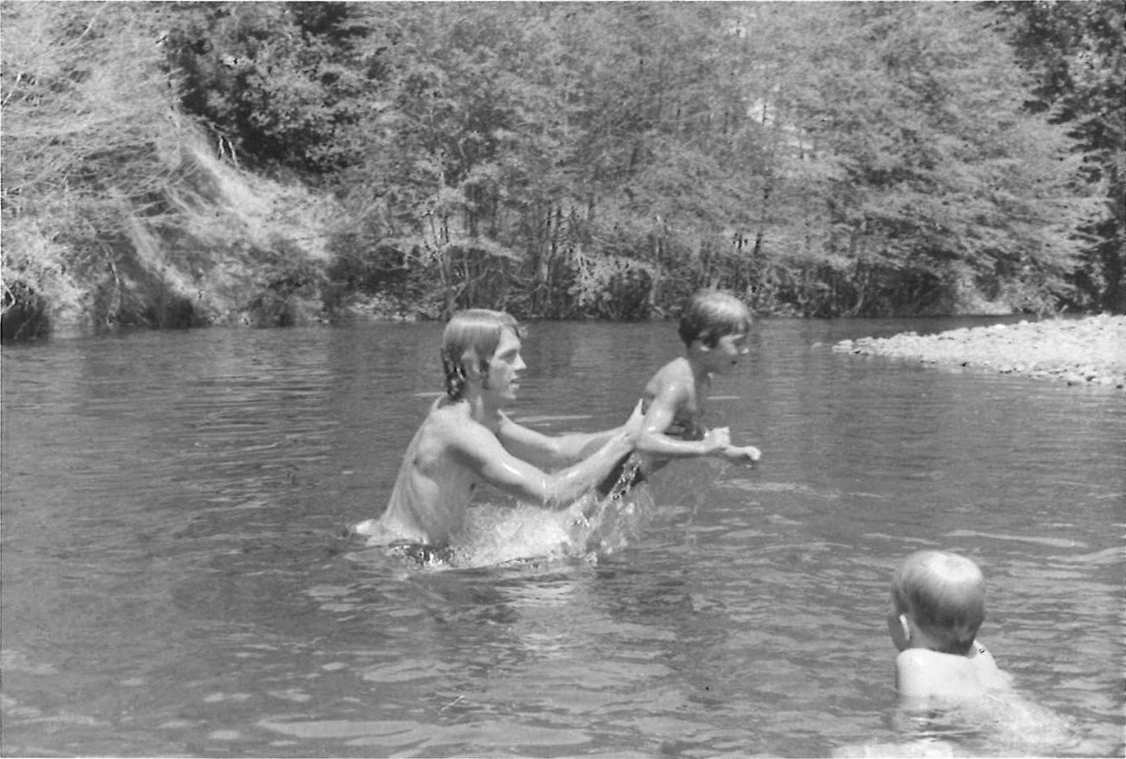 A person helps a child swim in a river, while another child watches. Background: trees and riverbank.