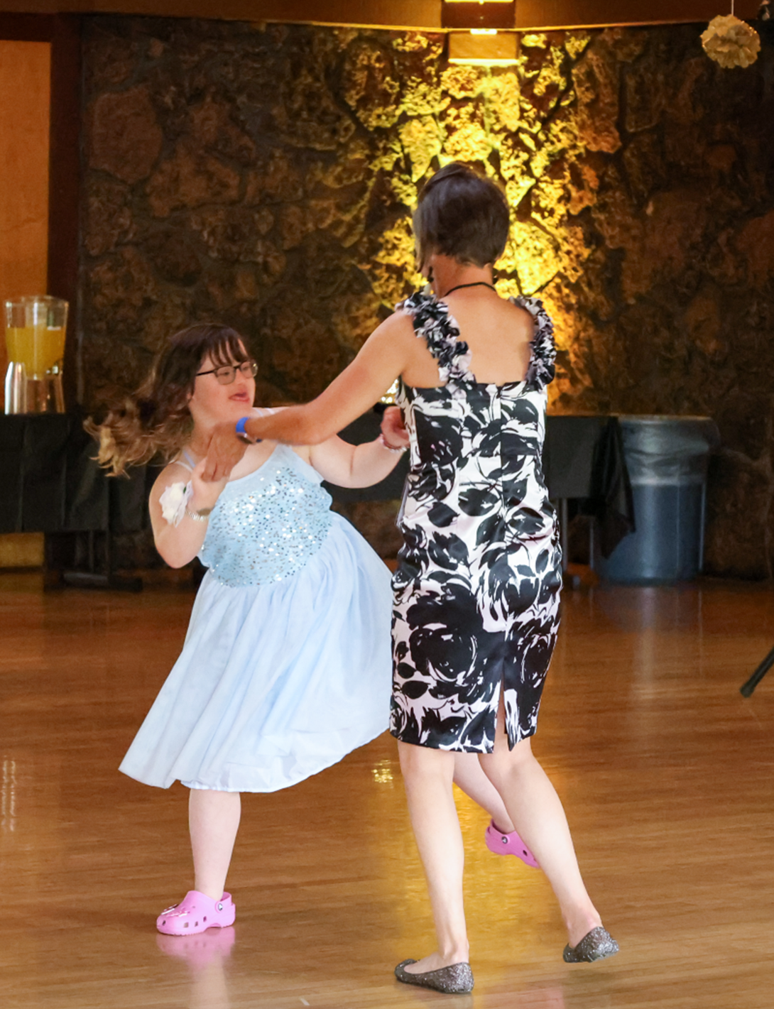 Two people dancing, one in a blue dress and one in a floral patterned dress, both wearing casual shoes.