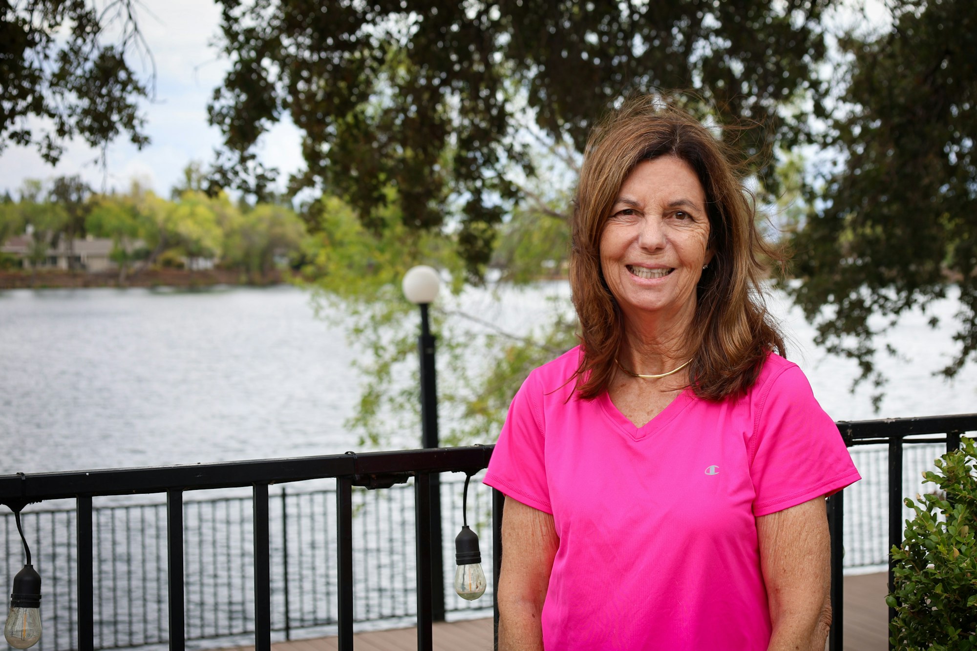 A woman in a pink shirt smiles by a lakeside with trees and a railing in the background.