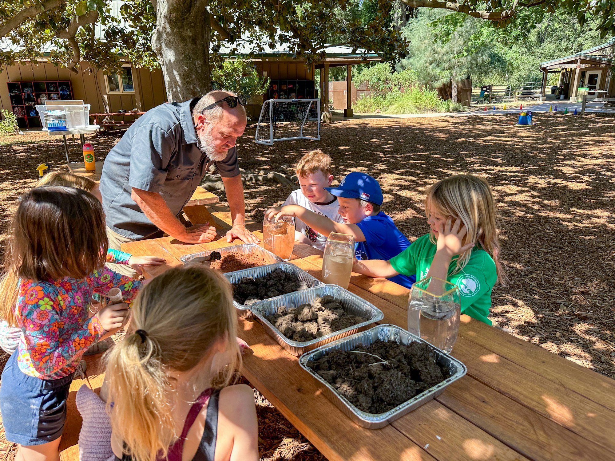 Children and an adult are gathered outdoors around a table with soil trays and jars of water, possibly for an educational activity.