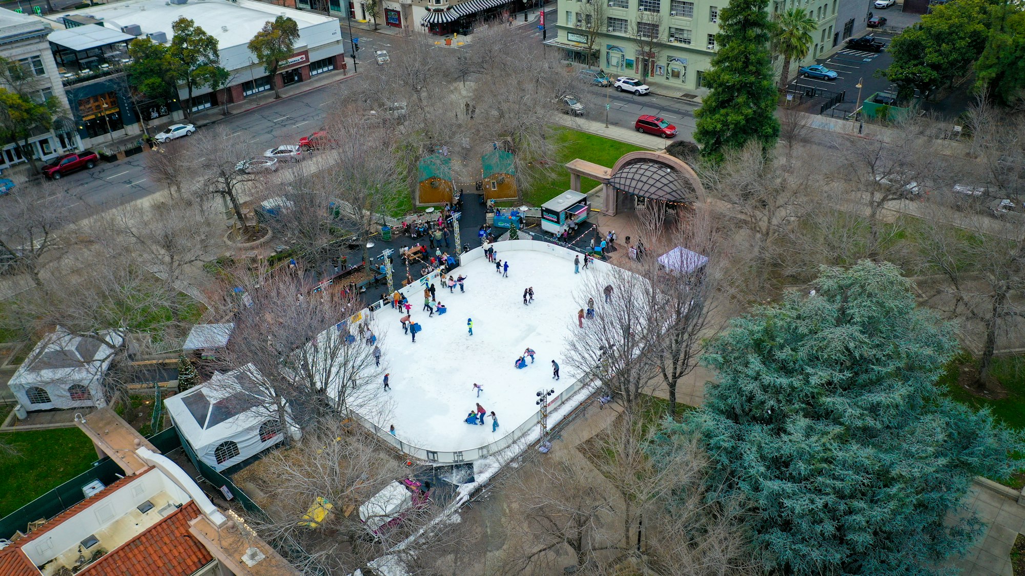Outdoor ice rink with people skating, surrounded by trees and buildings.