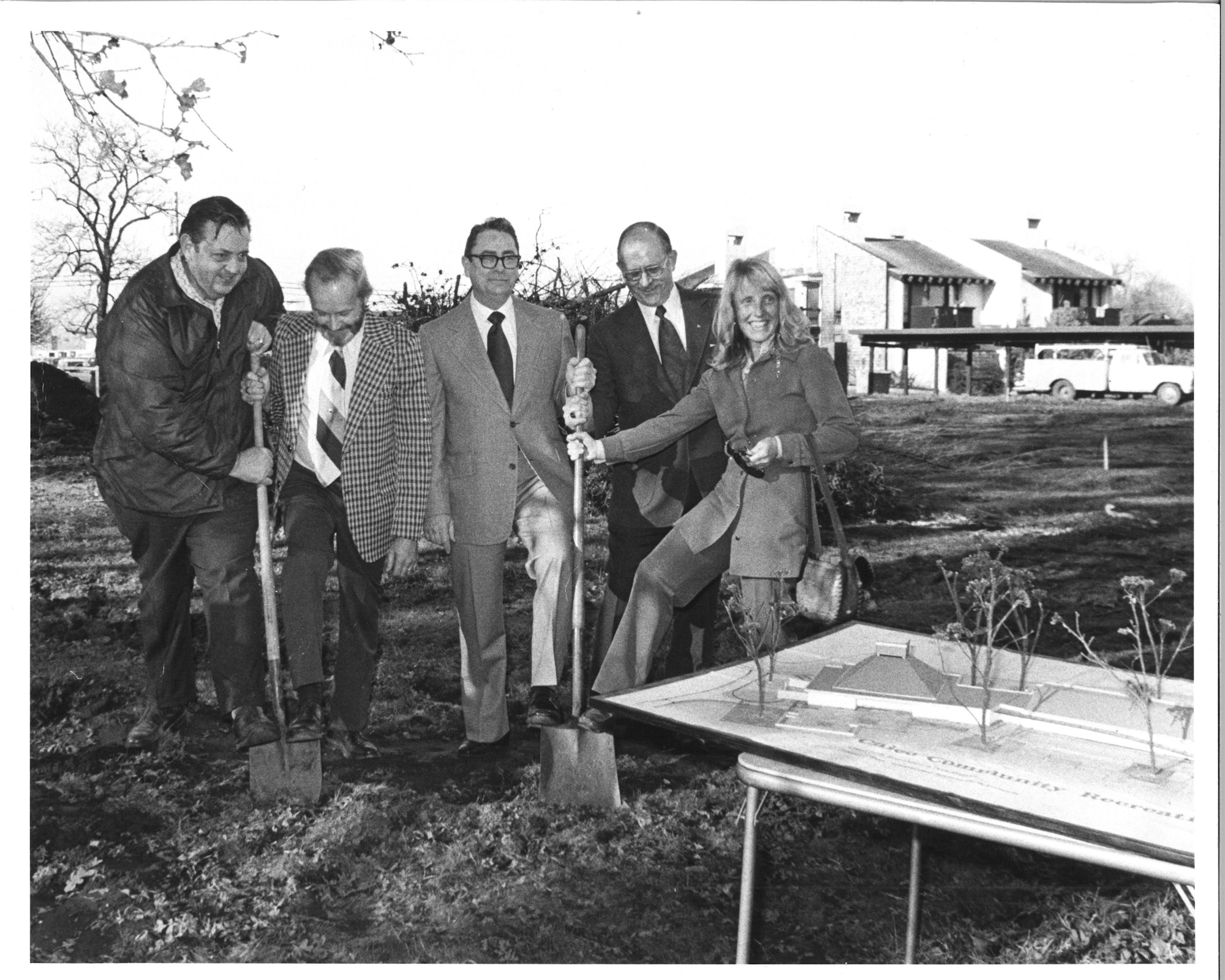 Five people in a groundbreaking ceremony with shovels, standing on a bare plot with a table displaying a building model.