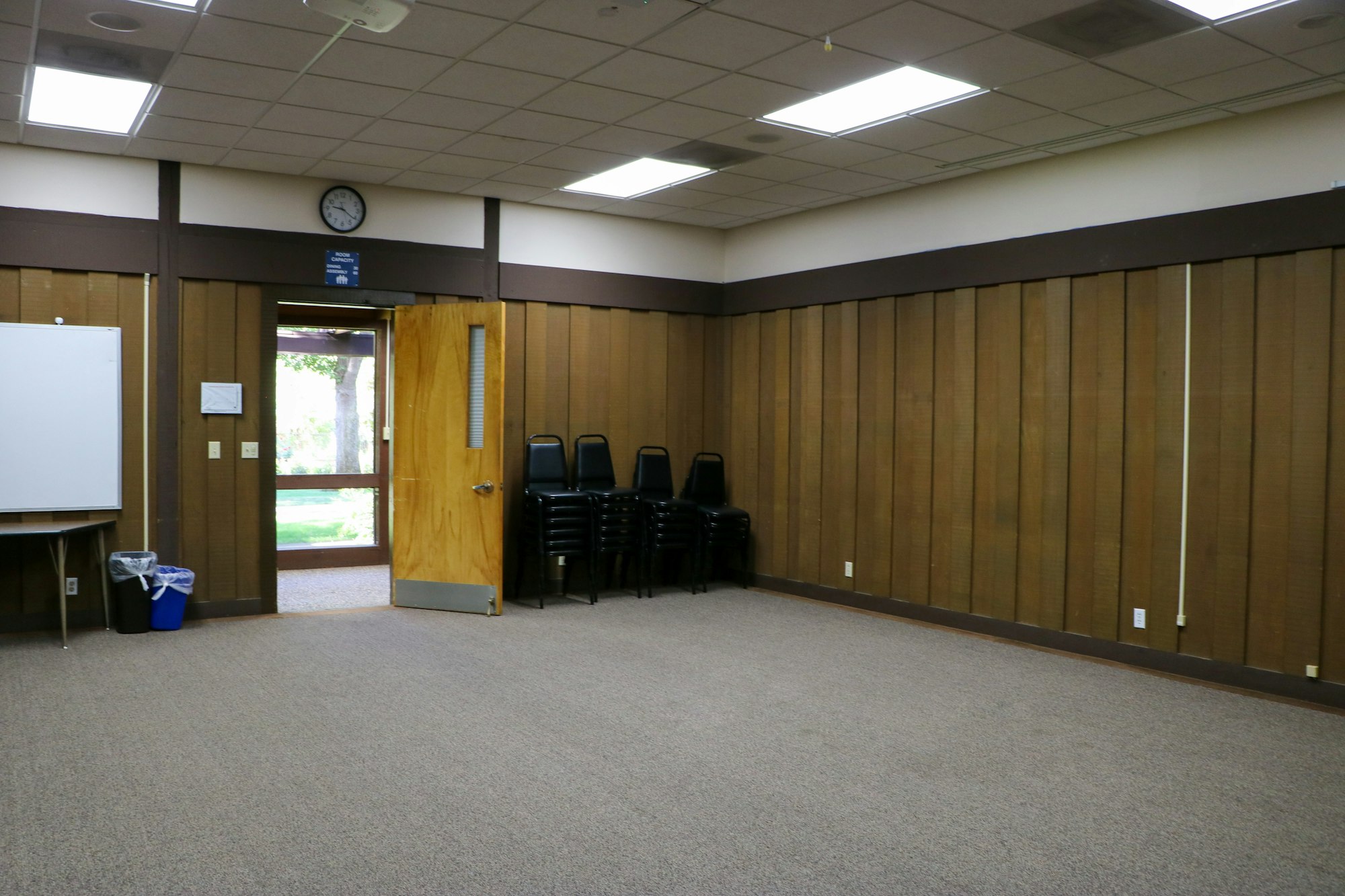 An empty meeting room with chairs stacked, a whiteboard, a clock on the wall, and a door open to the outside.