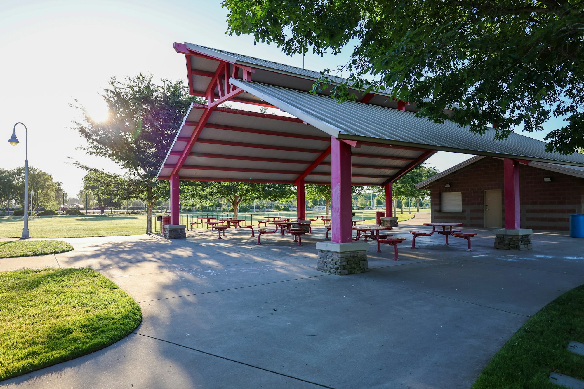 Sunlit park pavilion with picnic tables, grass, and trees.