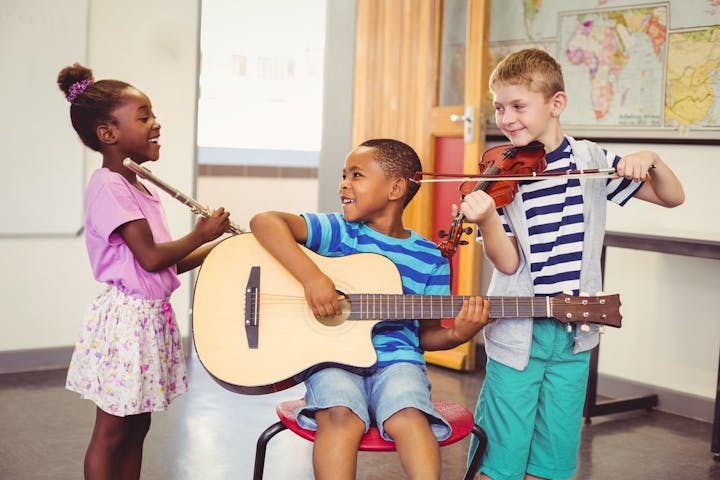 Three kids with a guitar, violin, and flute, smiling in a classroom.