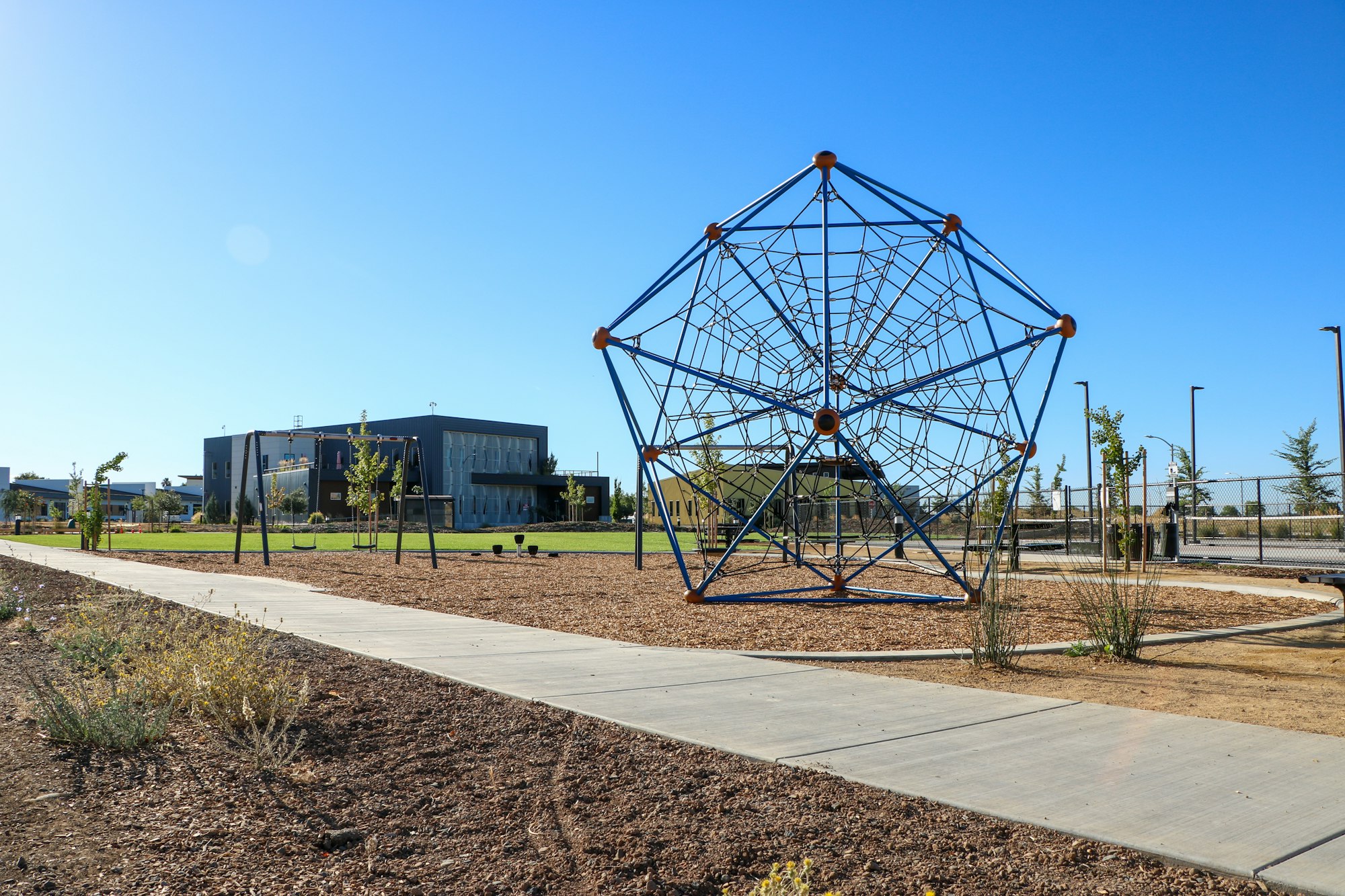 Climbing dome in a playground with blue sky, building in the background, and landscaped path in the foreground.