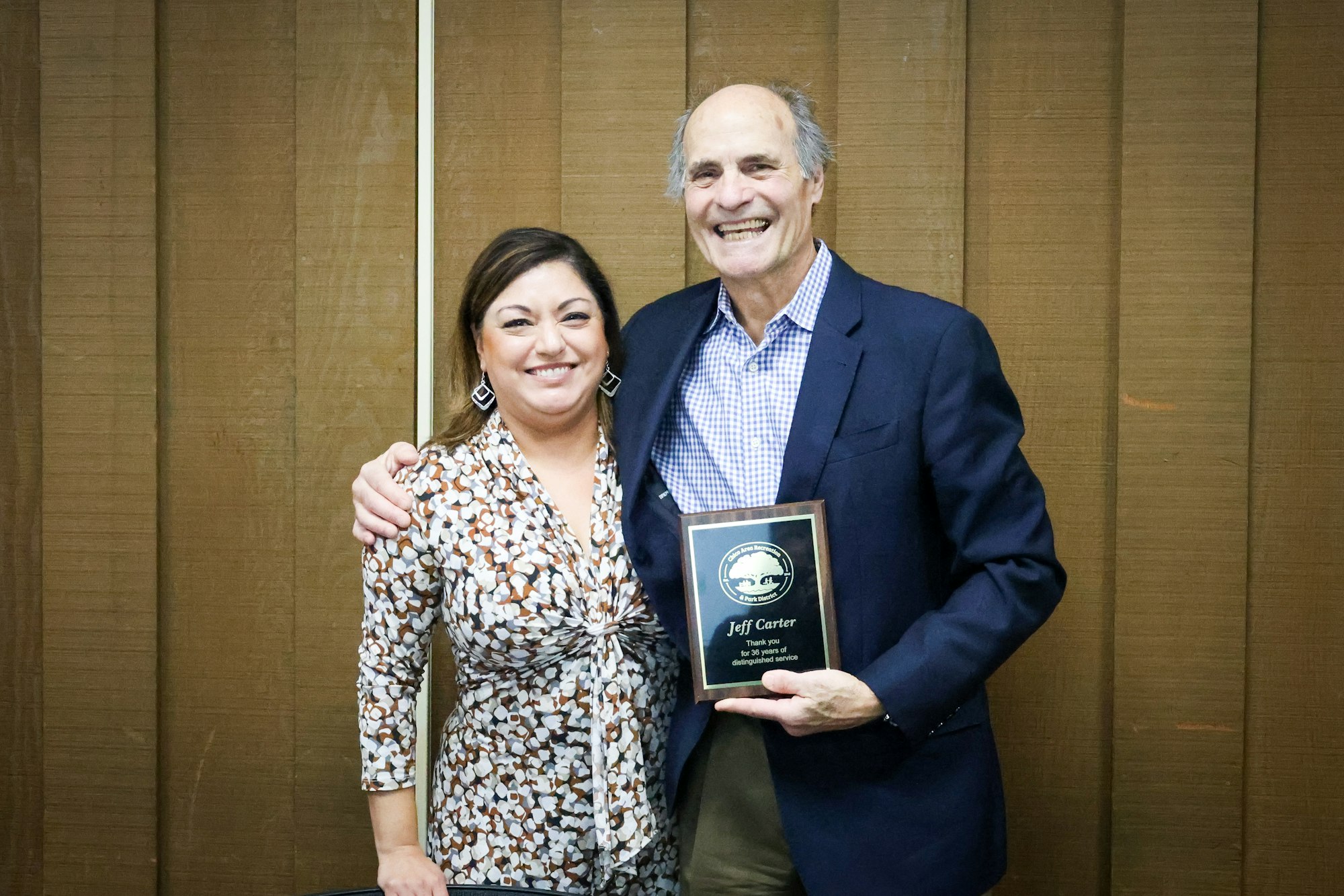Two people smiling, one holding an award plaque.