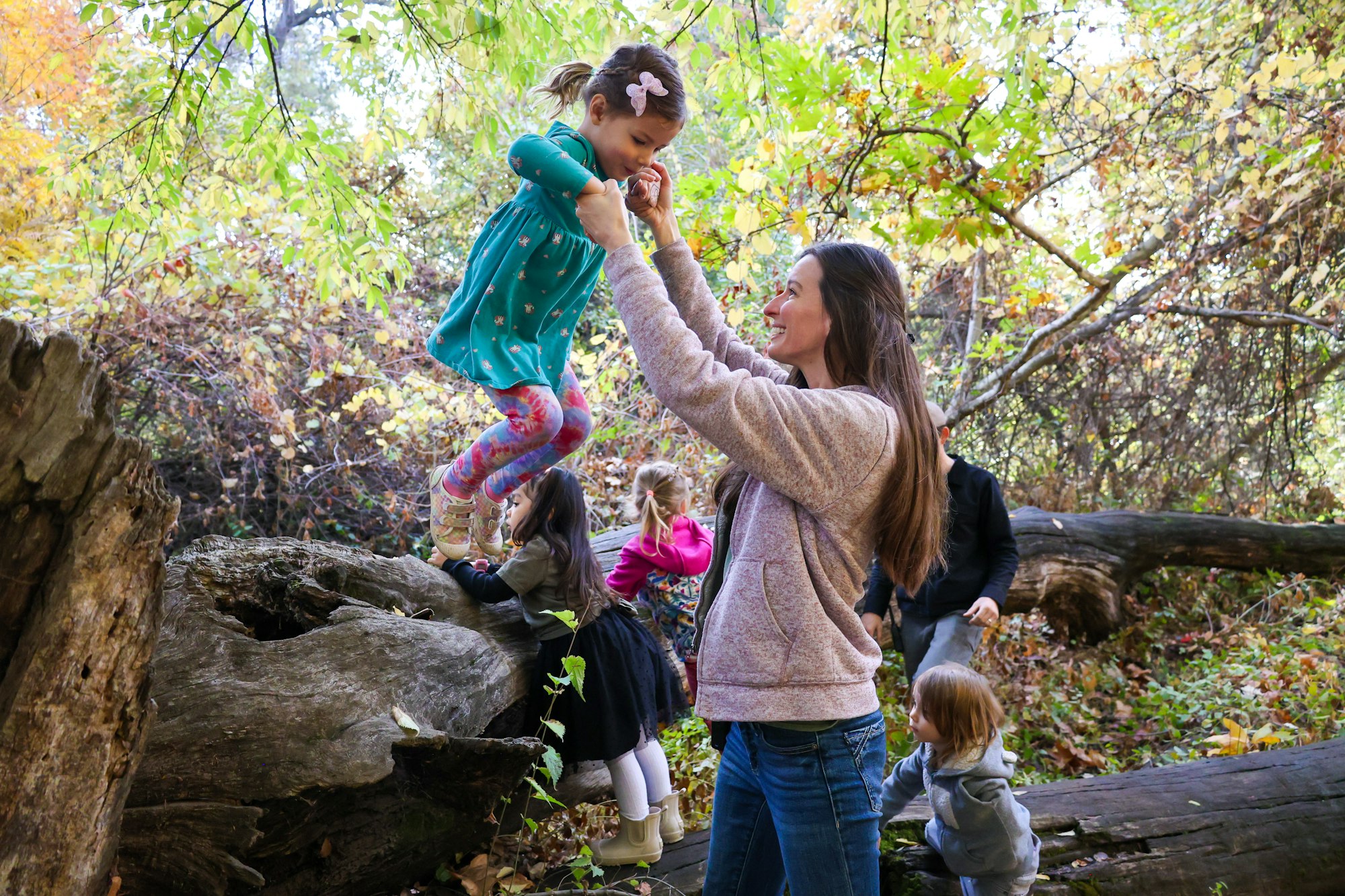 A woman helps a child jump from a log in a forested area, with other children playing around.