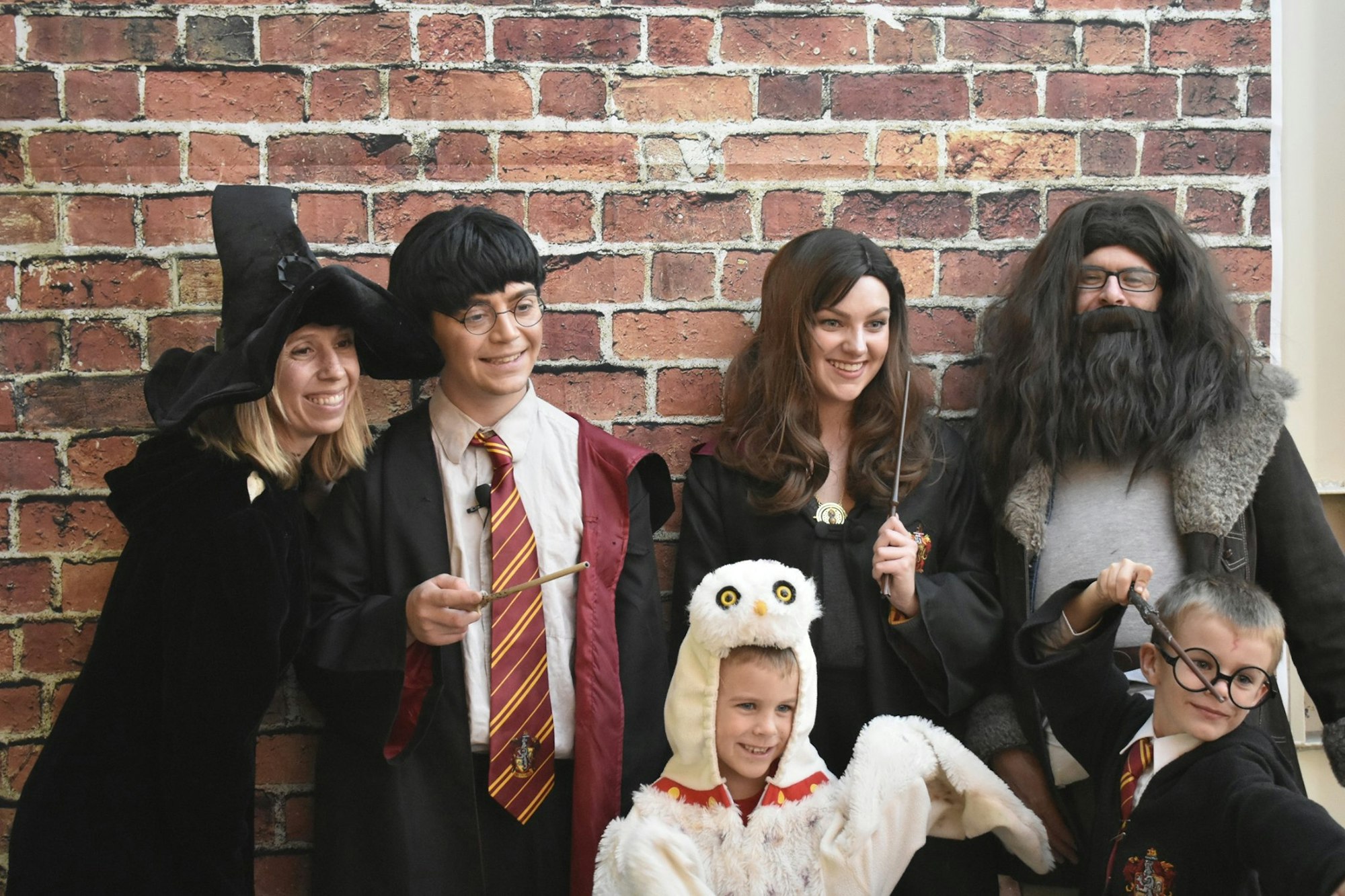 A group dressed as characters from a fantasy series poses in front of a brick wall, holding wands and wearing themed outfits.