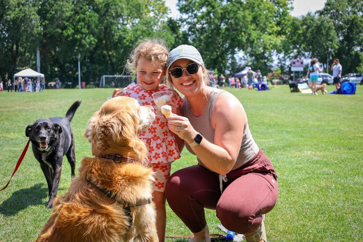 A smiling woman and a girl enjoy ice cream while two dogs play in a sunny park with people in the background.