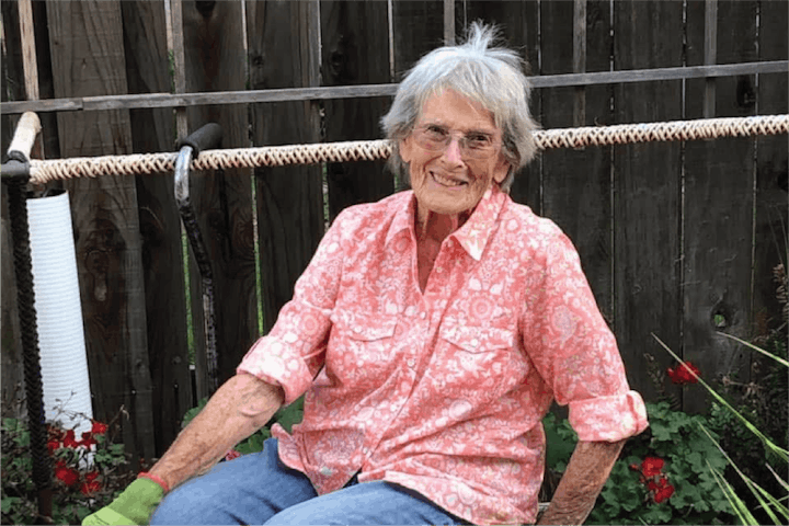 A person in a pink shirt sitting outdoors on a bench, smiling, with flowers and a wooden fence in the background.