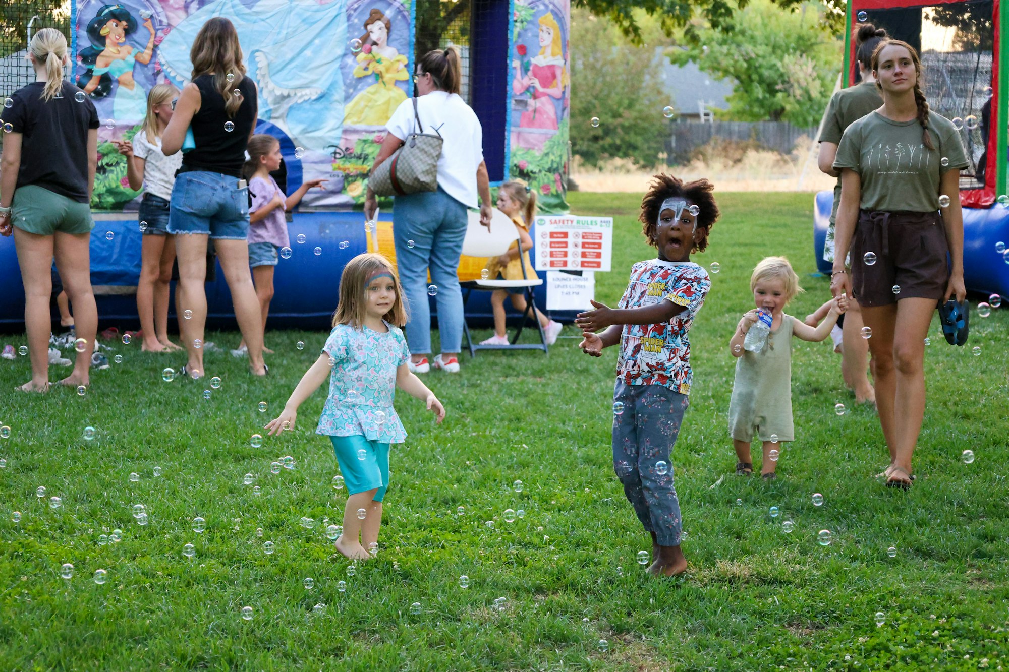 Children play among bubbles in a park, with adults nearby and a bouncy house in the background.