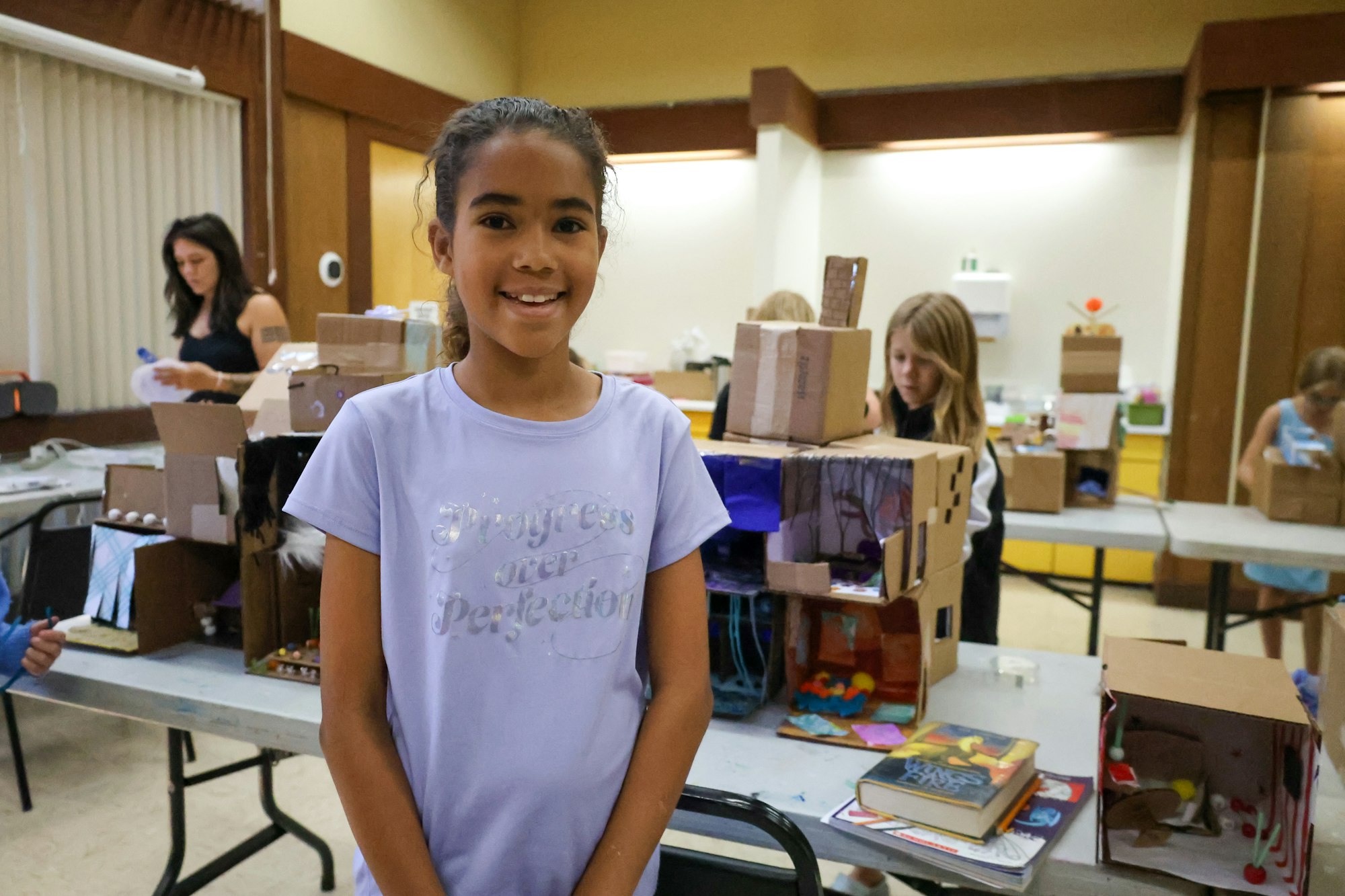 A girl in a classroom smiles, surrounded by cardboard projects, art supplies, and children working at tables.