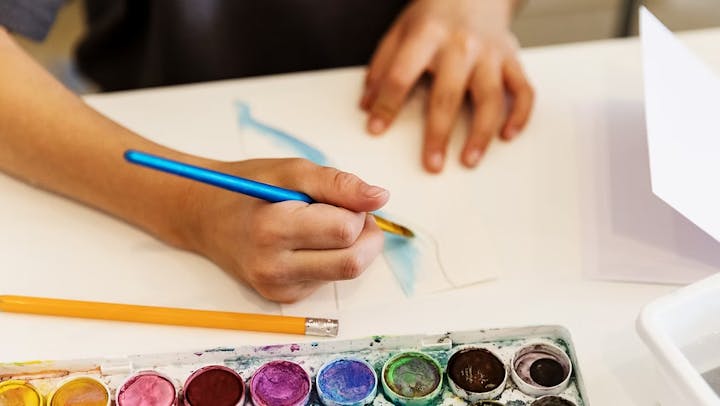 A person painting with watercolors, using a blue brush, next to a set of paints and a pencil on a white surface.