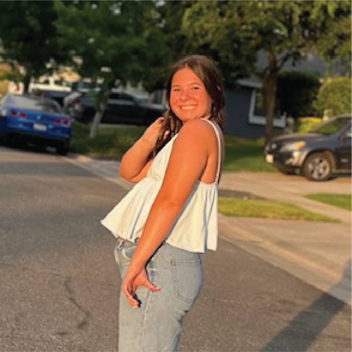 A person smiling, standing outside on a street, wearing a white top and blue jeans, with cars and trees in the background.