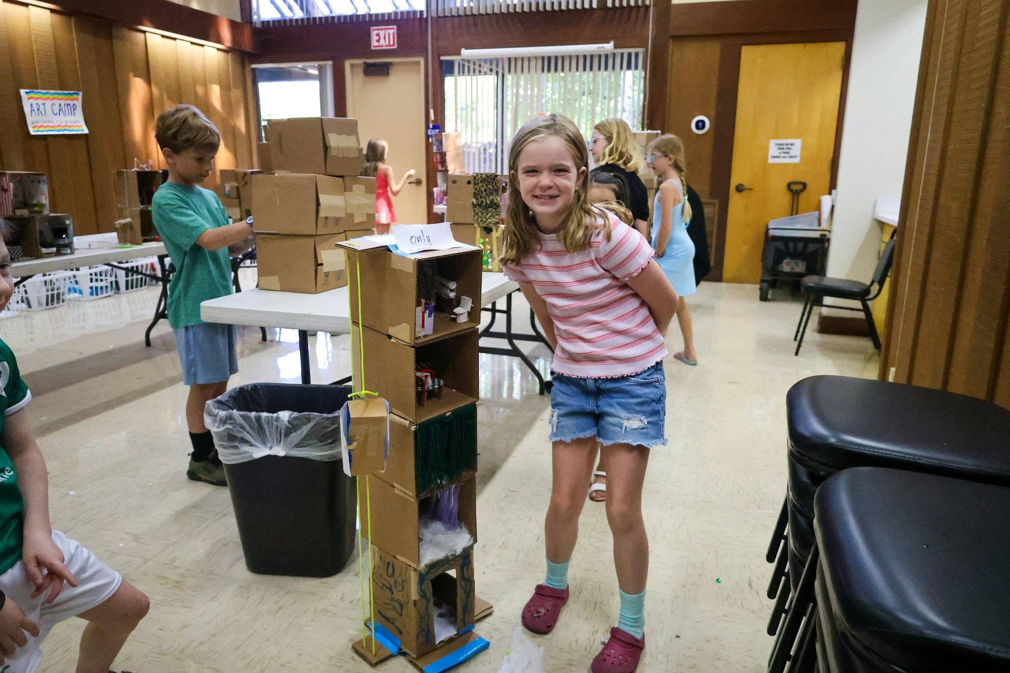Children at art camp with cardboard structures on display; a girl smiles next to a tall box sculpture.