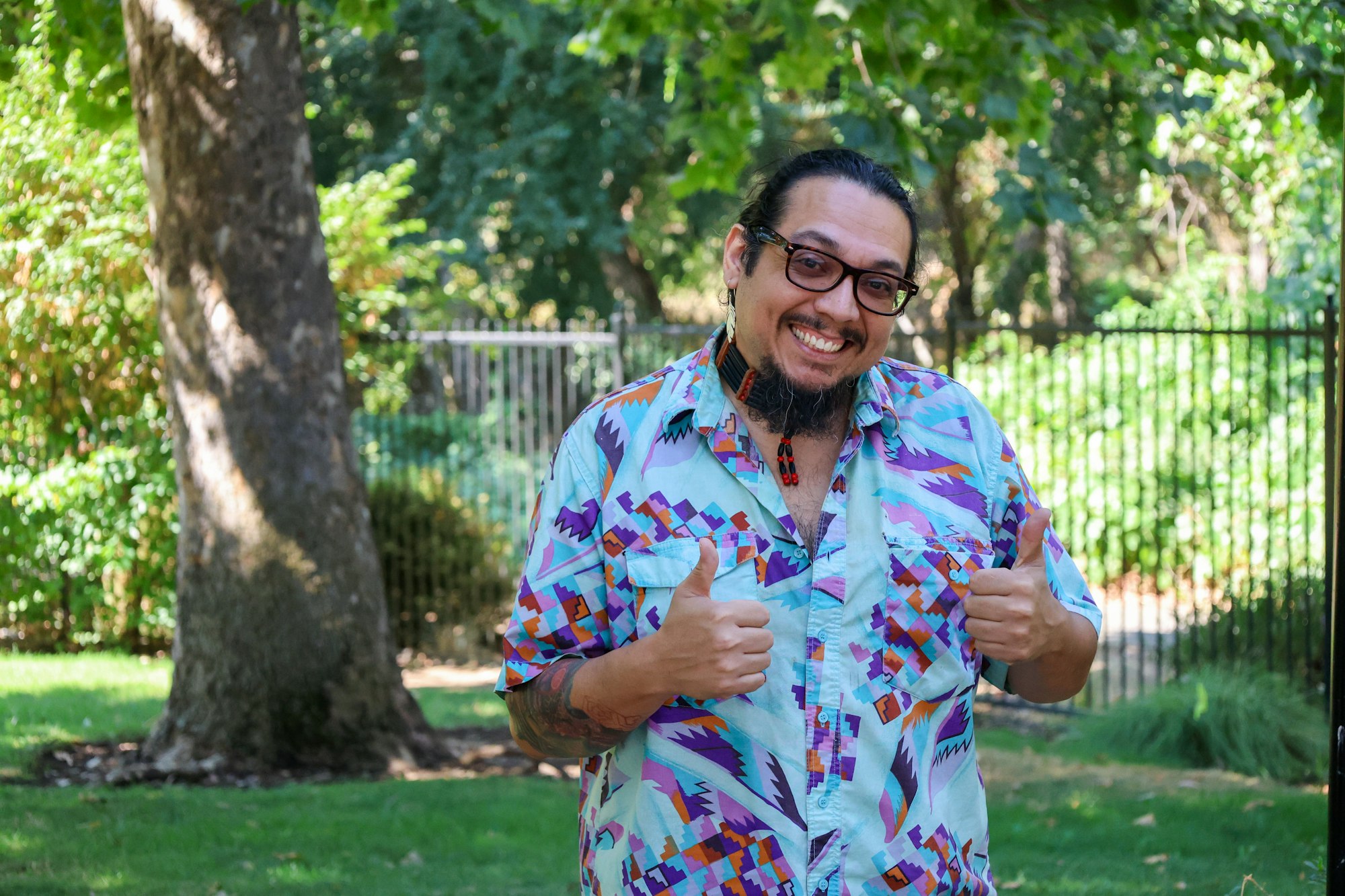 A smiling person giving thumbs up in a park with trees in the background.