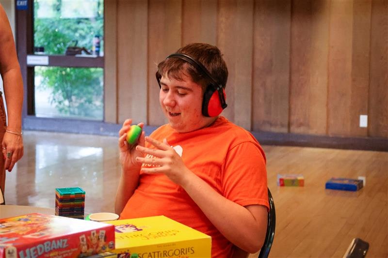 A person in an orange shirt and ear protectors holding a colorful object, with board games on a table nearby.