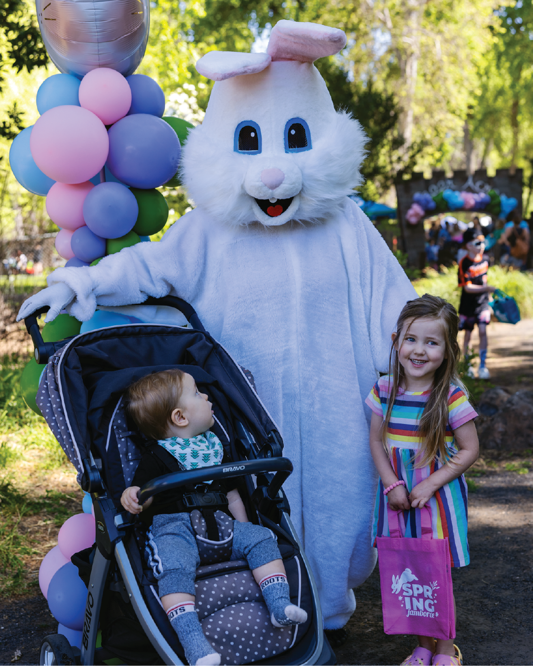 A costumed bunny stands with a young girl holding a bag, while a toddler sits in a stroller nearby, surrounded by colorful balloons.
