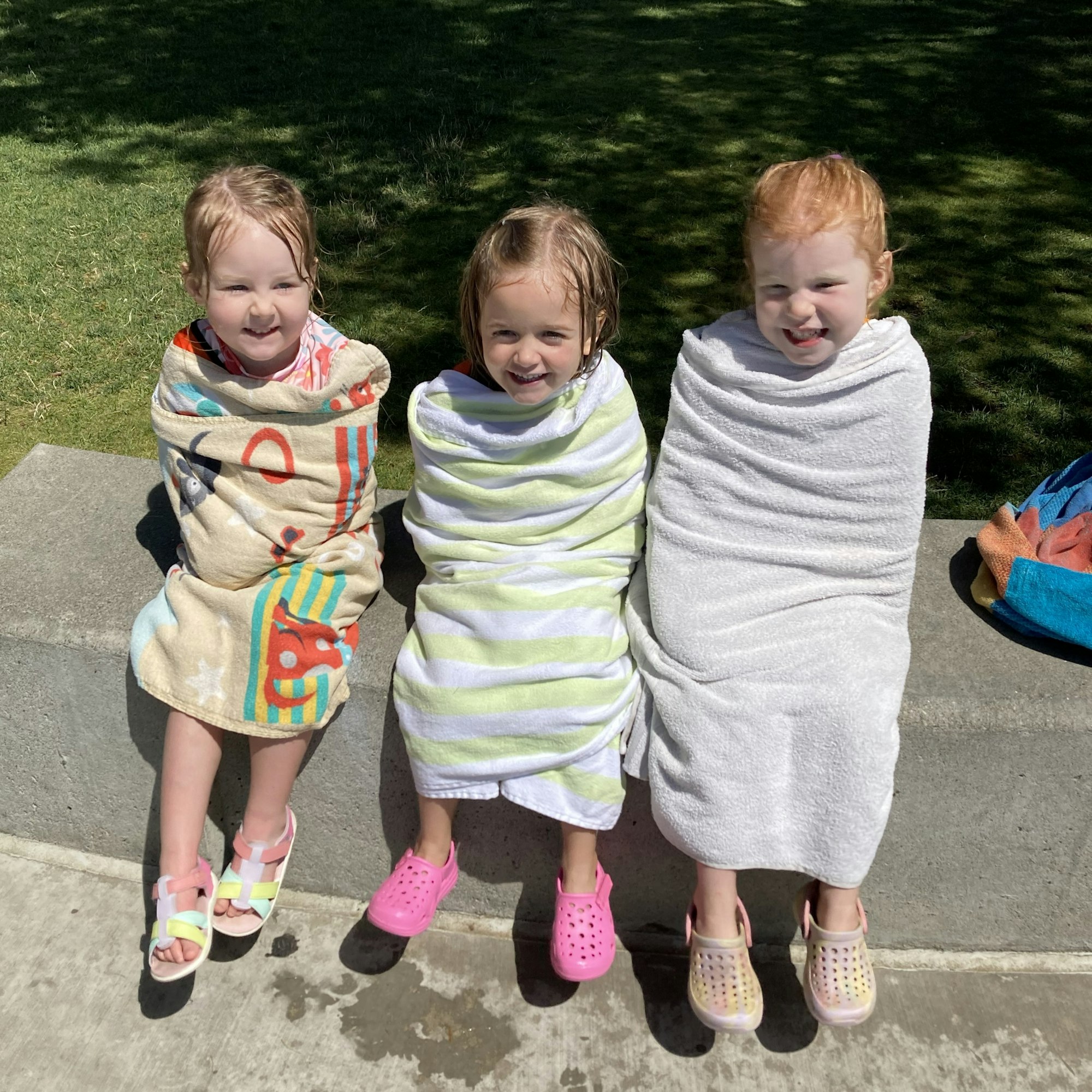Three young girls wrapped in towels sit on a ledge, smiling after swimming on a sunny day.